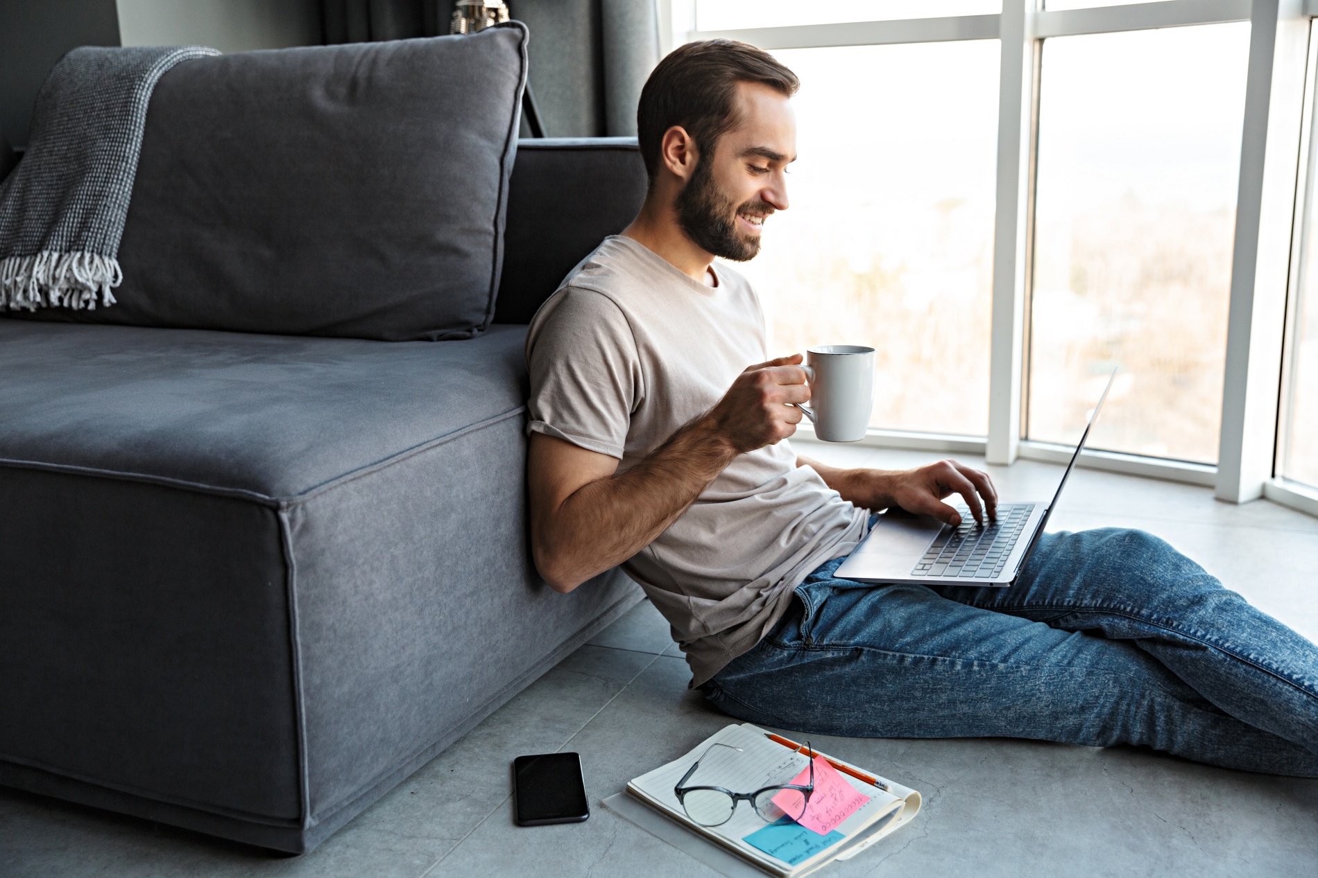 Attractive happy smart young man sitting on a floor in the living room, working on ;laptop computer, drinking coffee